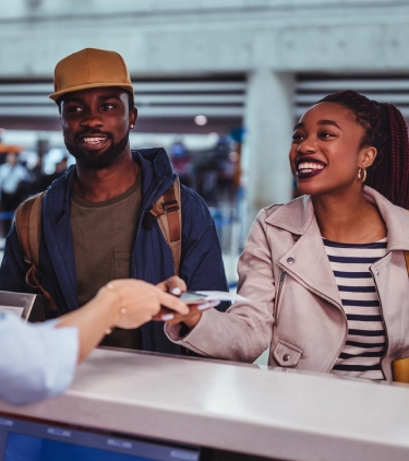 a young, black couple with couples' travel insurance speaks with an agent and hands over their boarding passes at the airport 