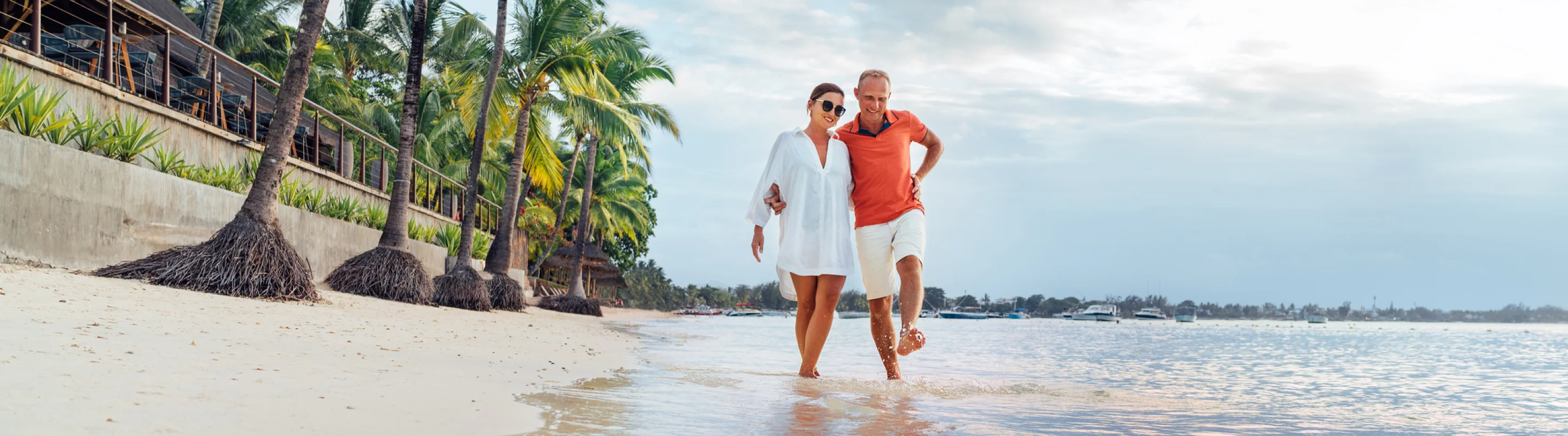 a couple with travel insurance for trip cancellations walks on a beach near the shoreline with palm trees in the background