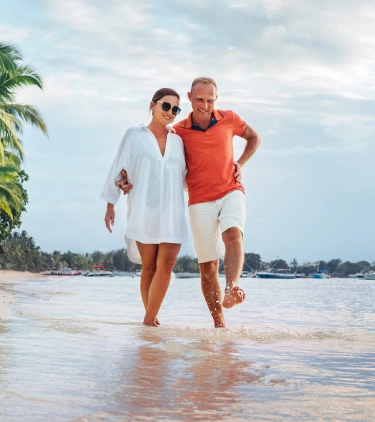 a couple with travel insurance for trip cancellations walks on a beach near the shoreline with palm trees in the background