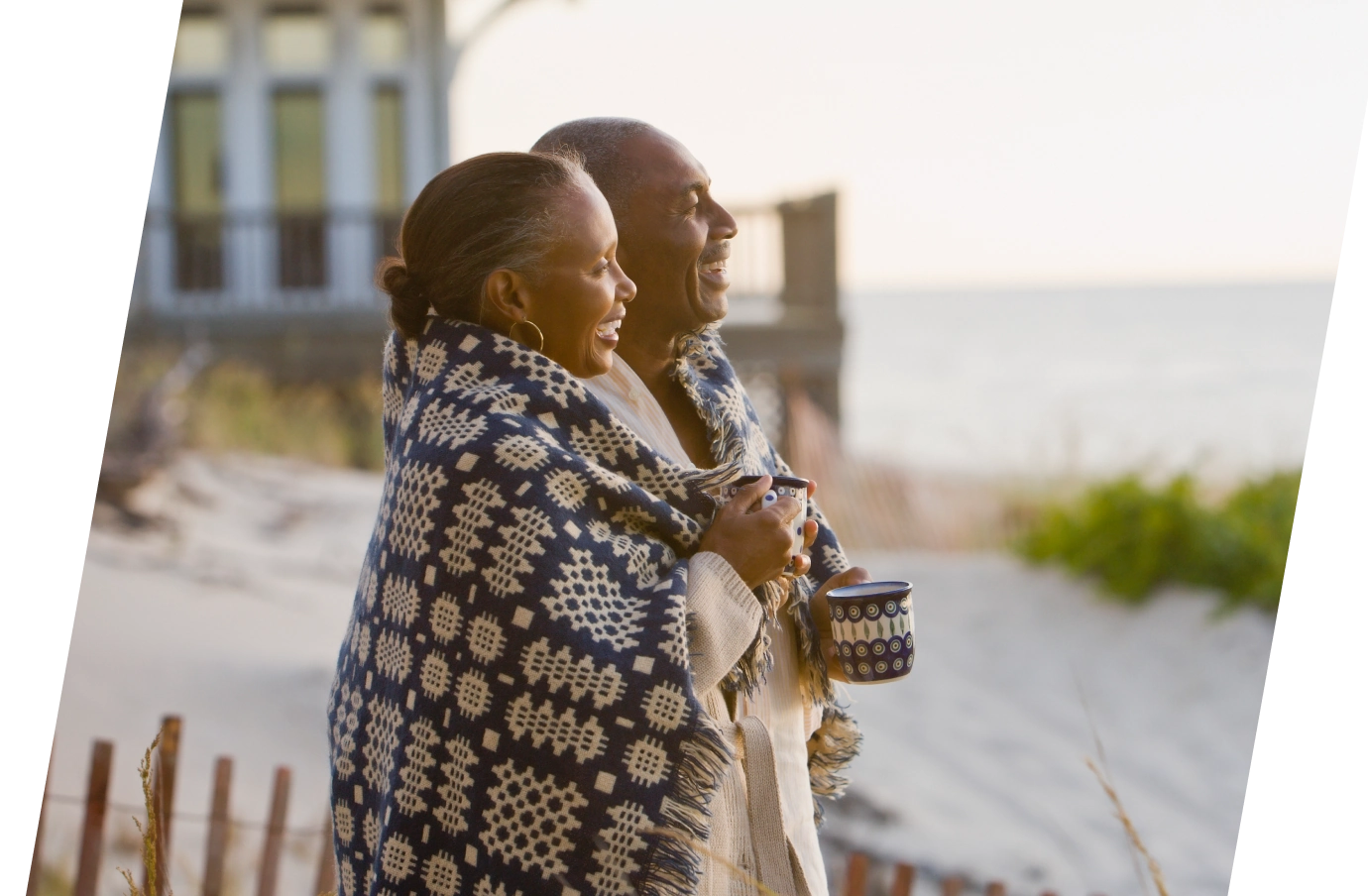 a middle-aged couple with couples' travel insurance holds mugs by the beach, wrapped in a blanket