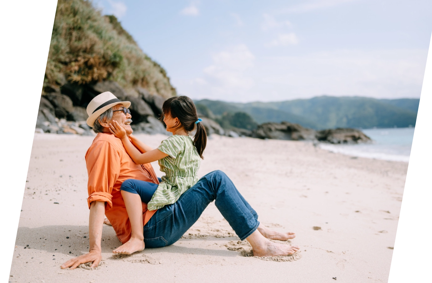 a grandfather with travel insurance for pre-existing conditions sits on a beach with his granddaughter near the shore 