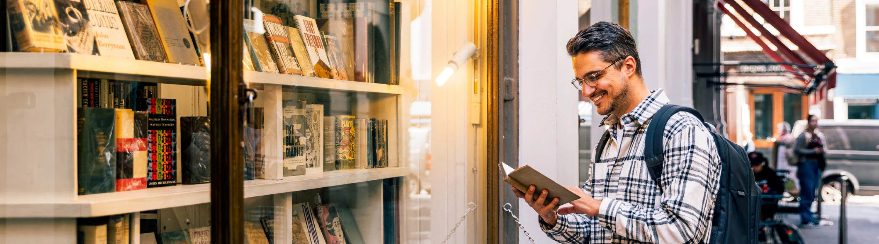 a young adult man with a backpack and travel insurance for students reads a book outside of a bookstore in a city