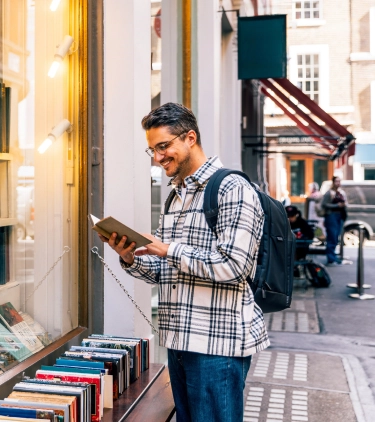 a young adult man with a backpack and travel insurance for students reads a book outside of a bookstore in a city
