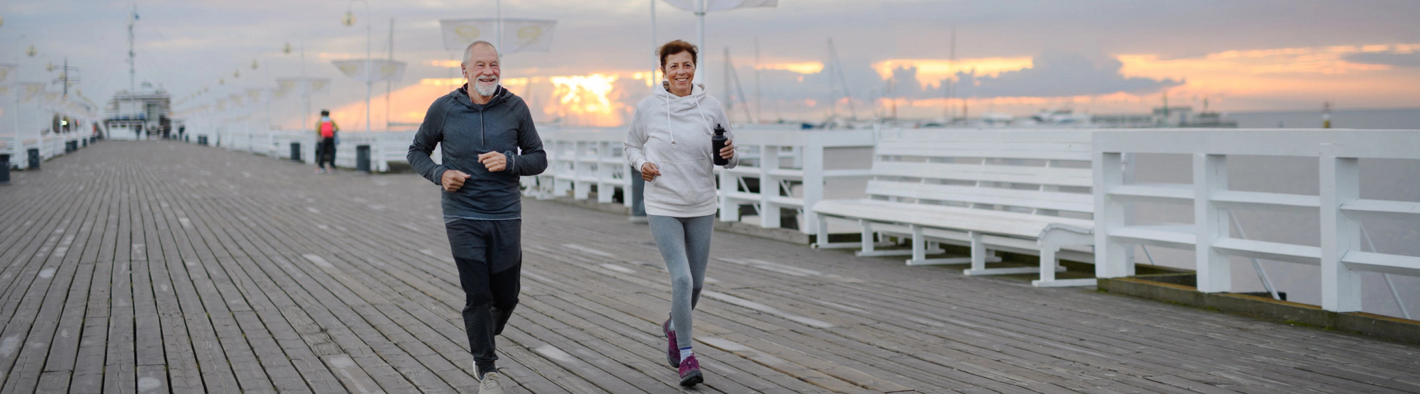 an older couple with travel insurance for missed connections jogs on a pier over the ocean at sunrise