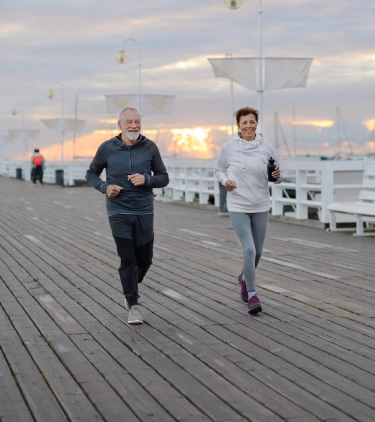 an older couple with travel insurance for missed connections jogs on a pier over the ocean at sunrise
