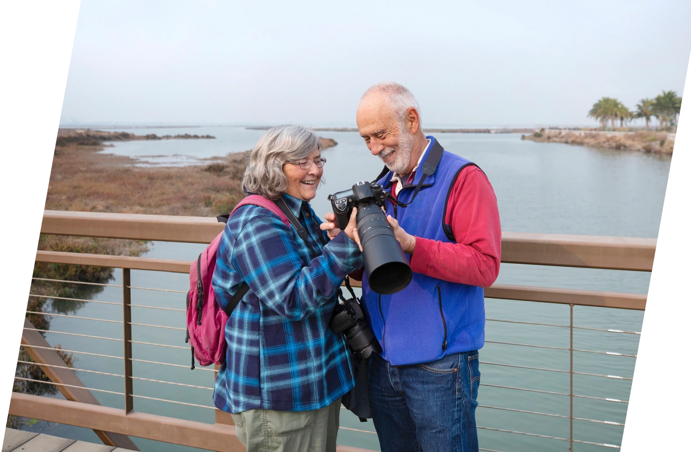 an older couple with travel insurance for pre-existing conditions reviews a camera’s photos on a wooden bridge over a marsh