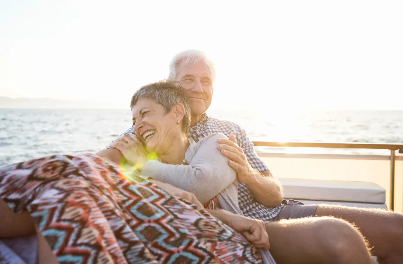 an older couple with emergency medical travel insurance embraces on a boat at sunset on vacation