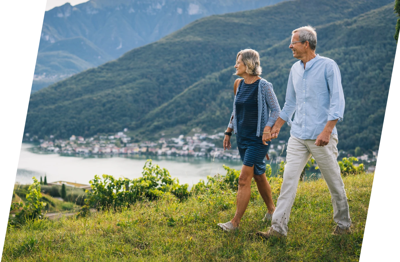 an older couple with travel insurance for medical evacuations walks hillside holding hands near a lake and town
