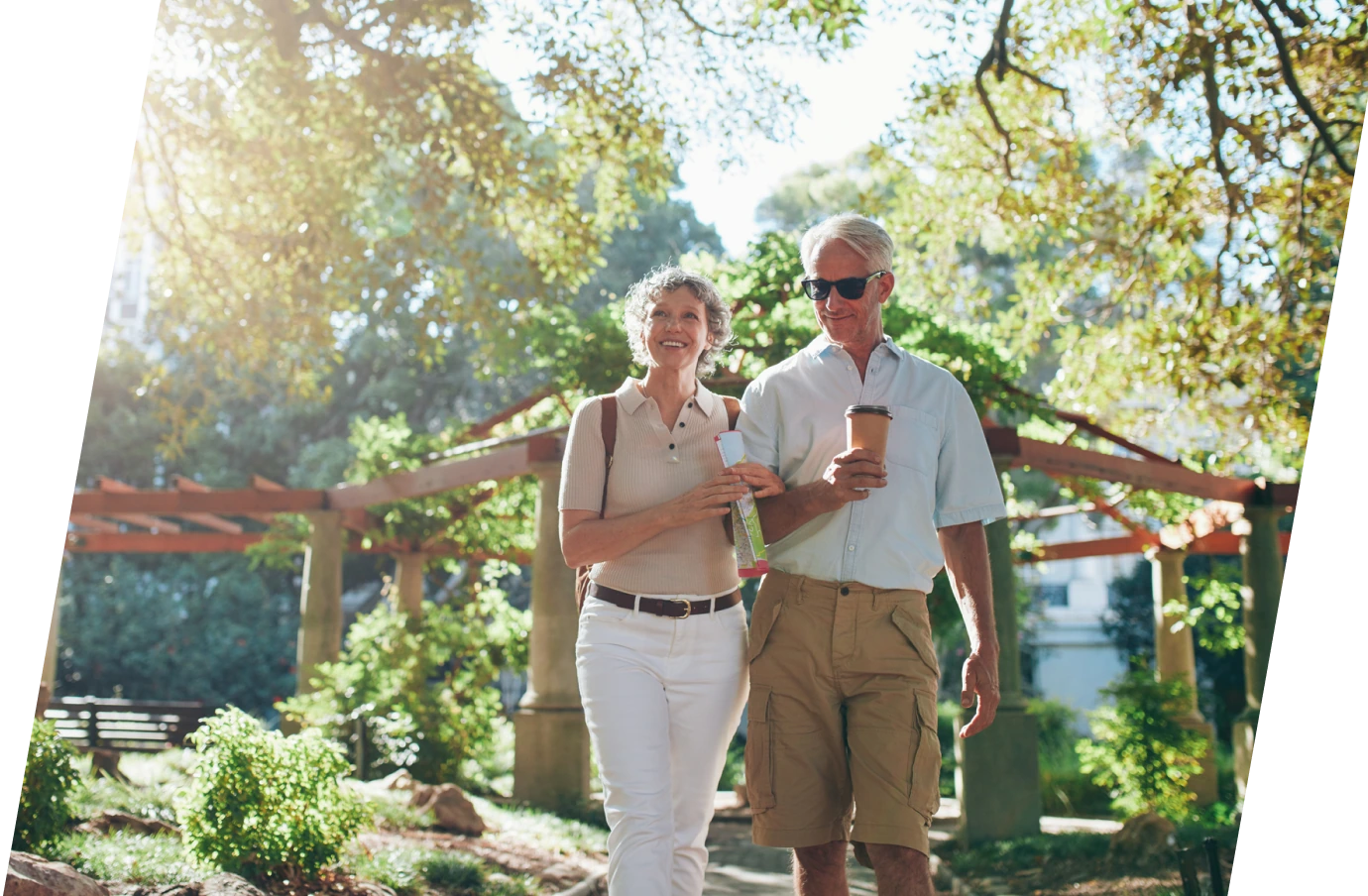 a senior couple with travel assistance enjoy a leisurely walk through a garden in a park holding a pamphlet and a to-go coffee