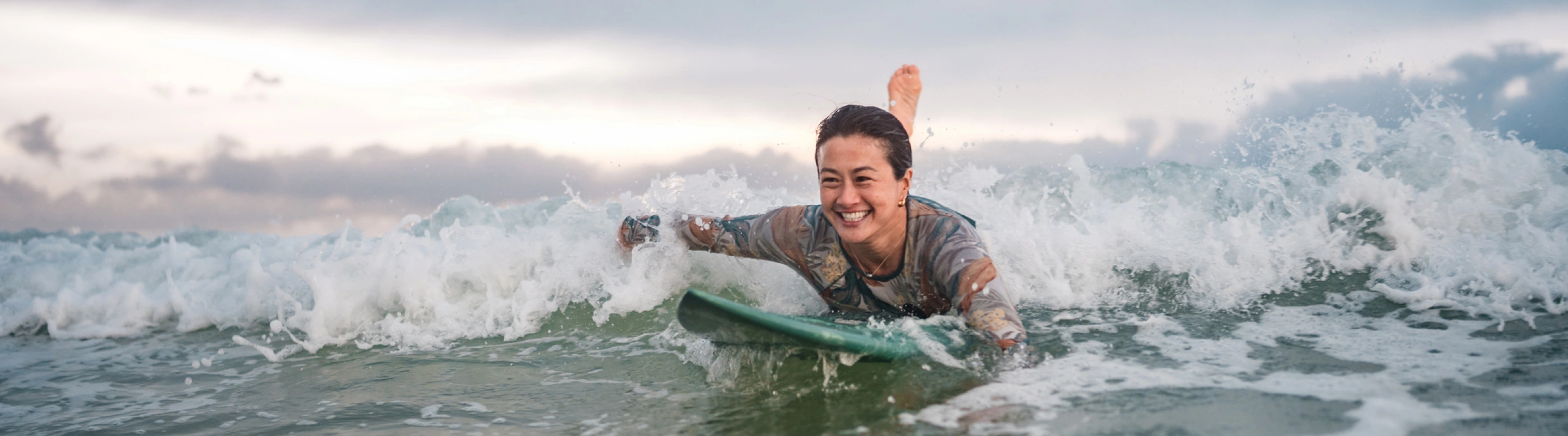 a female surfer with emergency medical insurance paddles through the ocean in a wetsuit on a cloudy day 