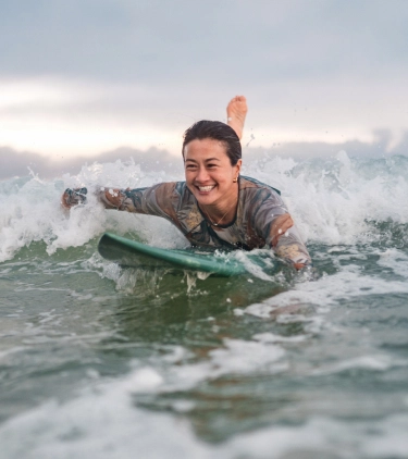 a female surfer with emergency medical insurance paddles through the ocean in a wetsuit on a cloudy day 