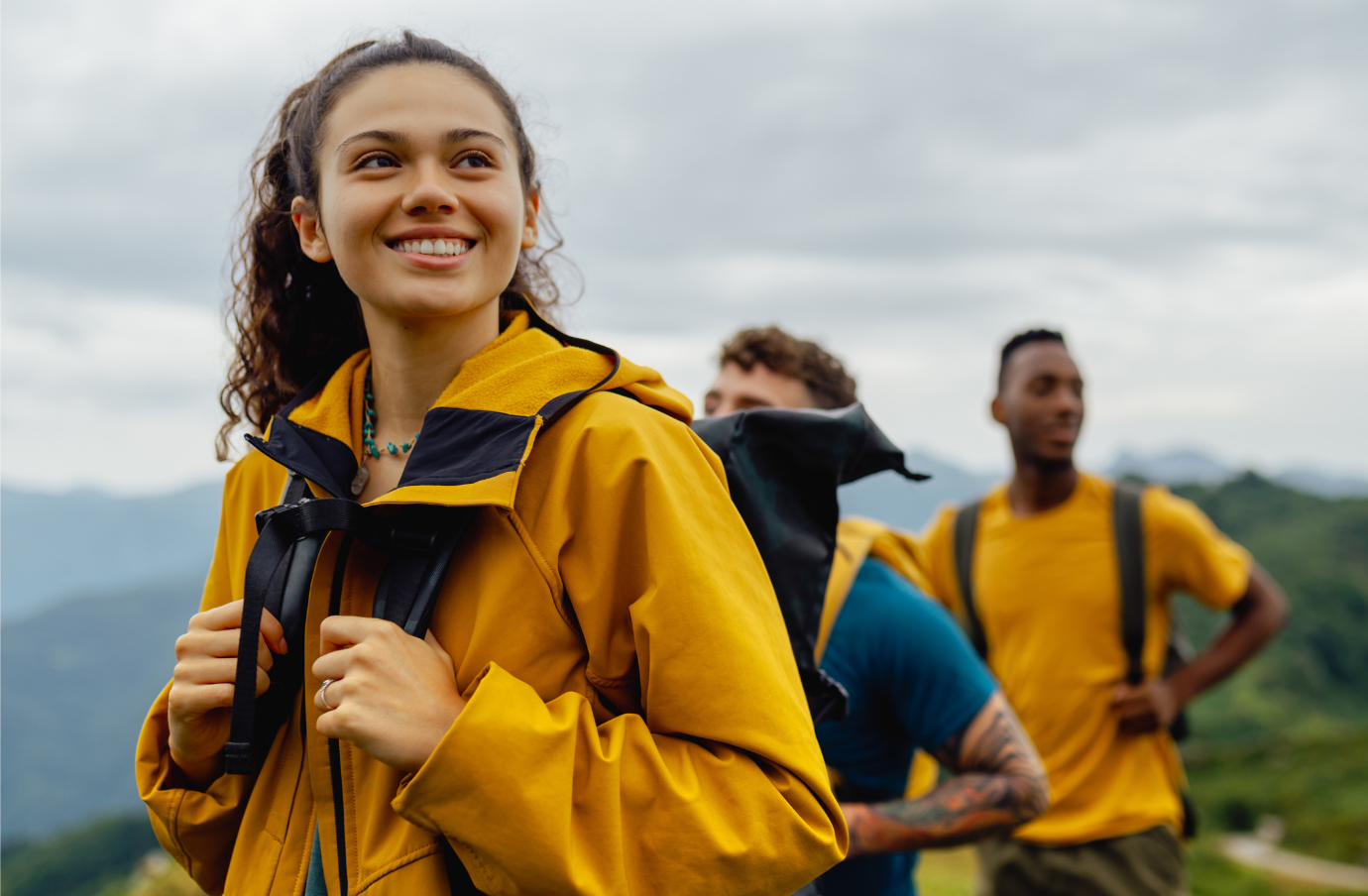 three young adults, with travel insurance for students and yellow hiking gear, smile as they hike a mountain trail