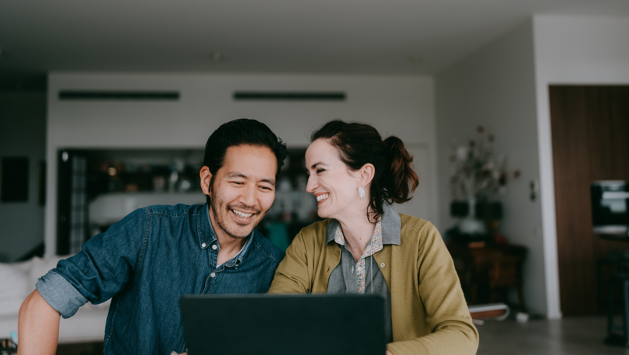 a couple smiles in front of a laptop, looking up how to calculate trip cost for travel insurance purposes