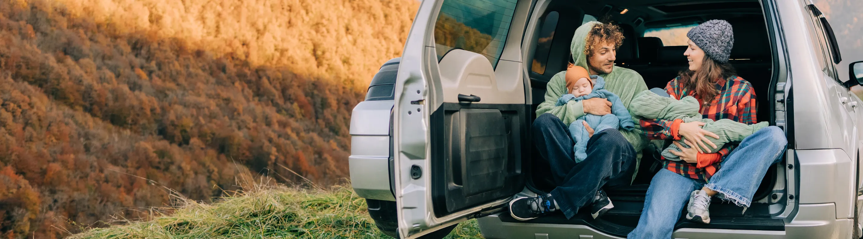 a mother and father with road trip travel insurance sit on the back of their SUV holding babies in the mountains  