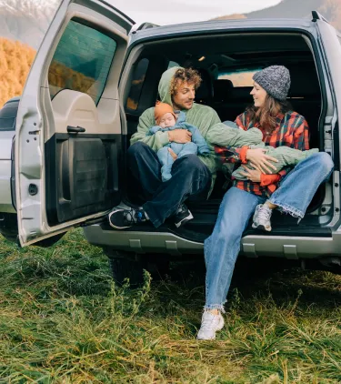 a mother and father with road trip travel insurance sit on the back of their SUV holding babies in the mountains  