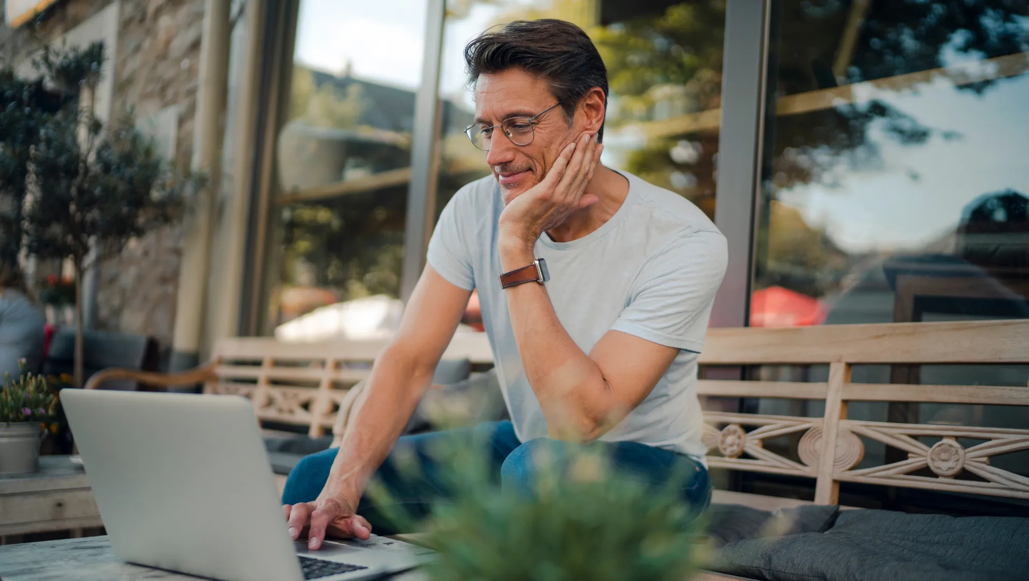a man looks at his laptop on an outdoor patio, researching how to calculate his trip cost for travel insurance purposes