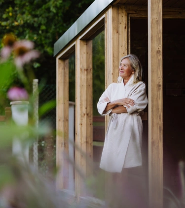 an older woman with travel insurance for wellness retreats stands outside a wooden spa in her robe