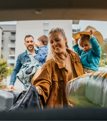 two young parents with family travel insurance unpack baggage from their SUV on vacation while holding their children