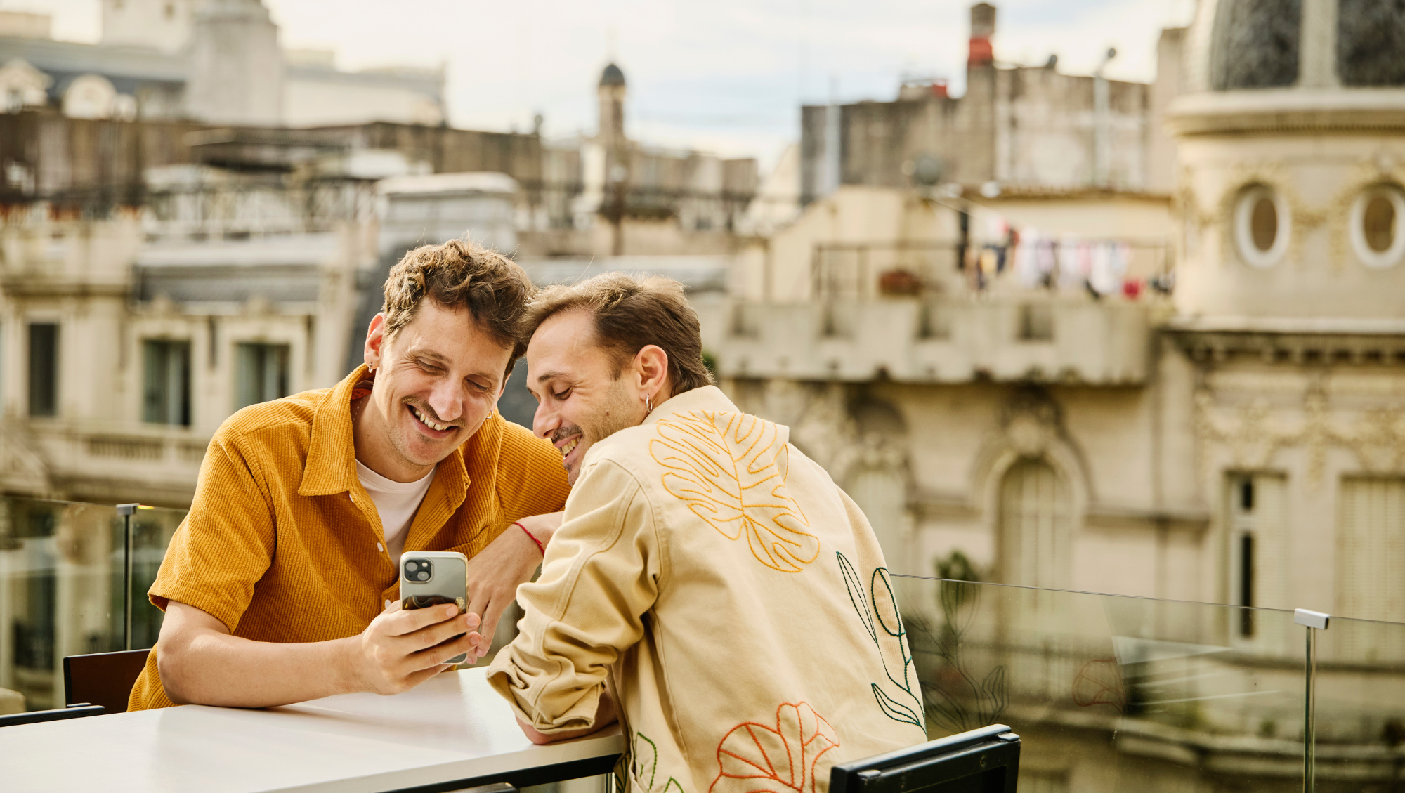 two partners sit on a European-looking rooftop, looking at the travel app on their phone for safety notifications