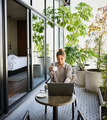 a remote worker with business travel insurance sits on a hotel balcony with a coffee and a laptop