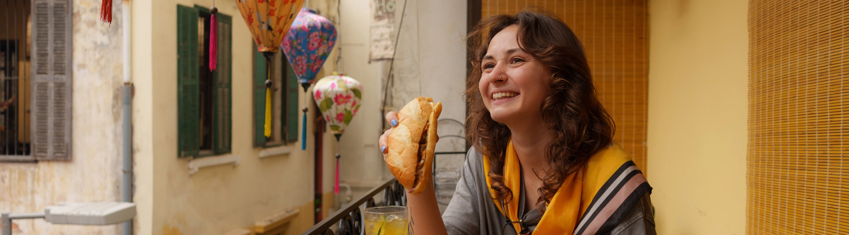 a young woman with travel insurance for culinary trips eats a baguette on a balcony near Vietnamese lanterns 
