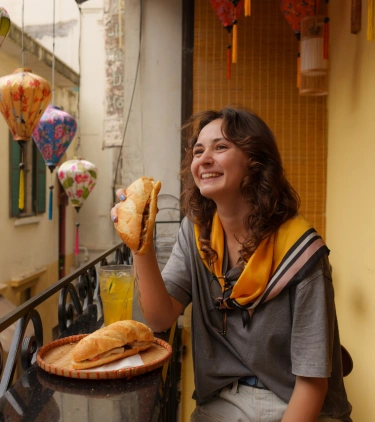 a young woman with travel insurance for culinary trips eats a baguette on a balcony near Vietnamese lanterns 
