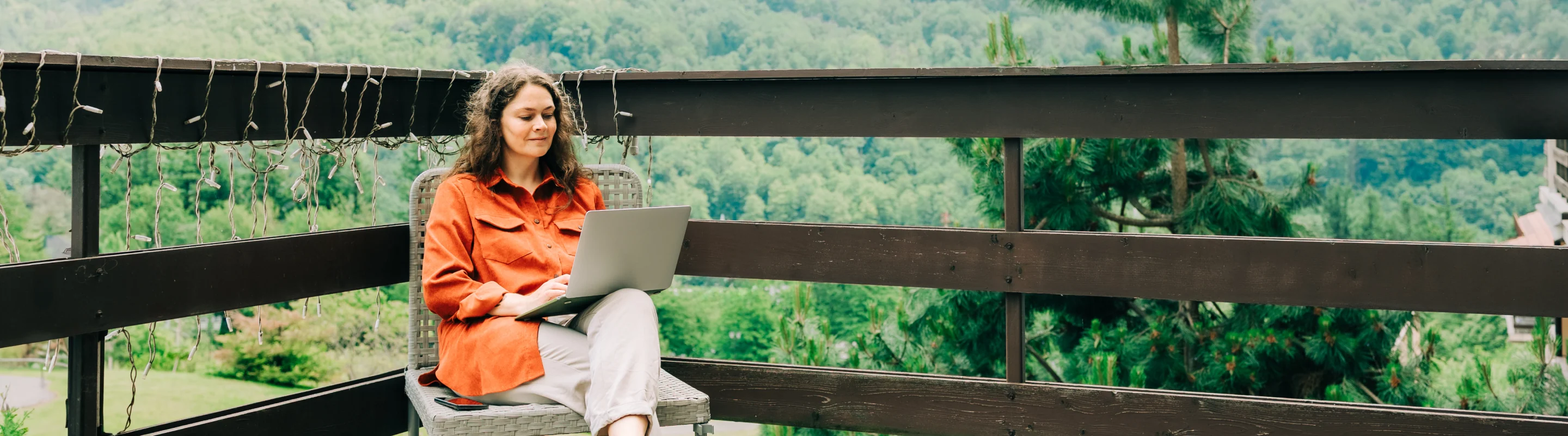 Young woman with travel insurance for digital nomads using her laptop on a deck with mountains in the background