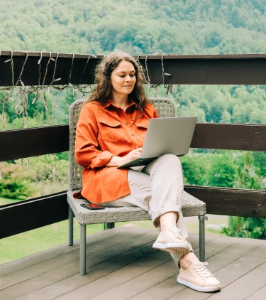 Young woman with travel insurance for digital nomads using her laptop on a deck with mountains in the background