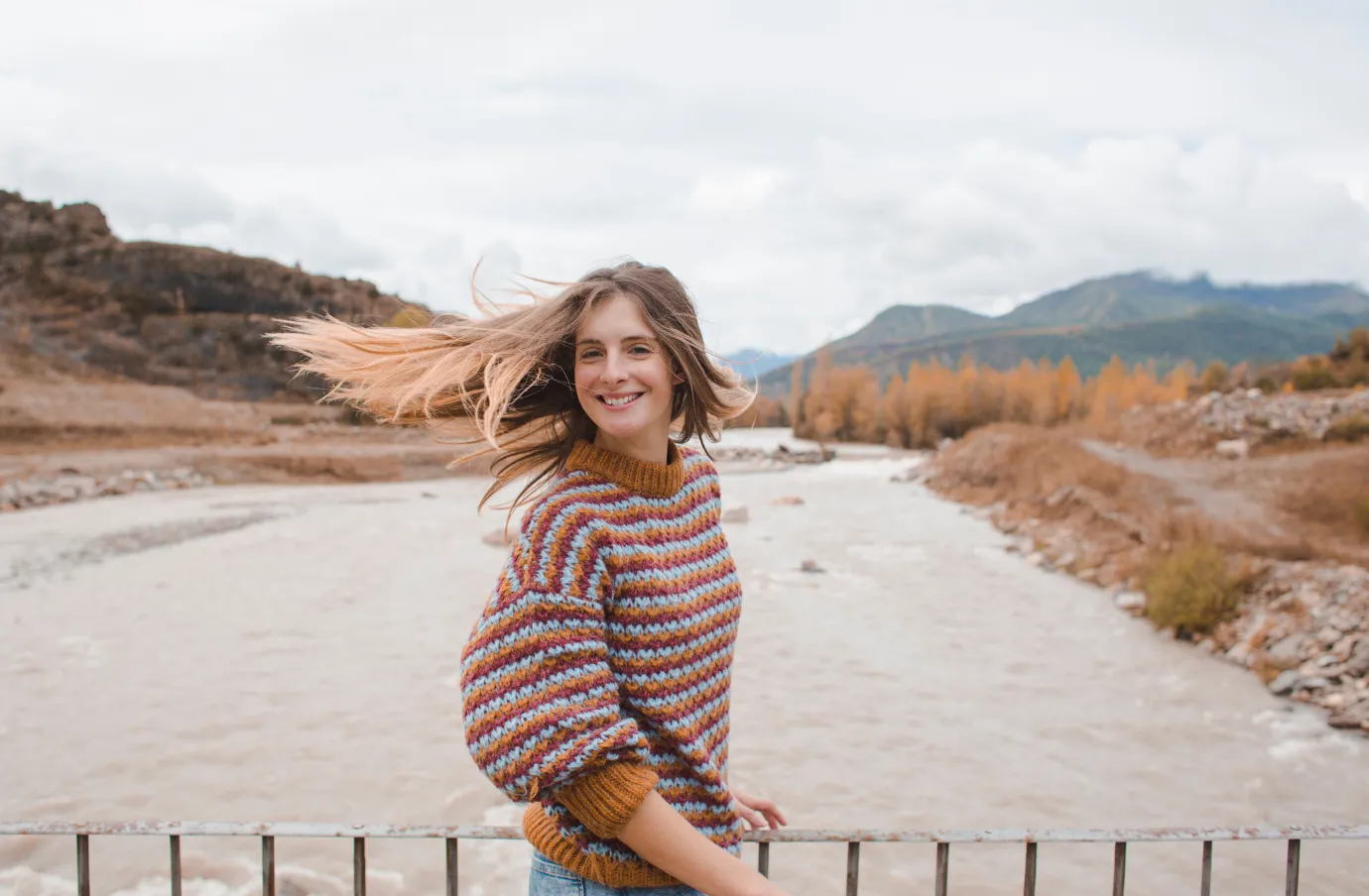 a woman flips her hair and smiles on a river bridge, surrounded by mountains and insured with travel insurance for nature trips 