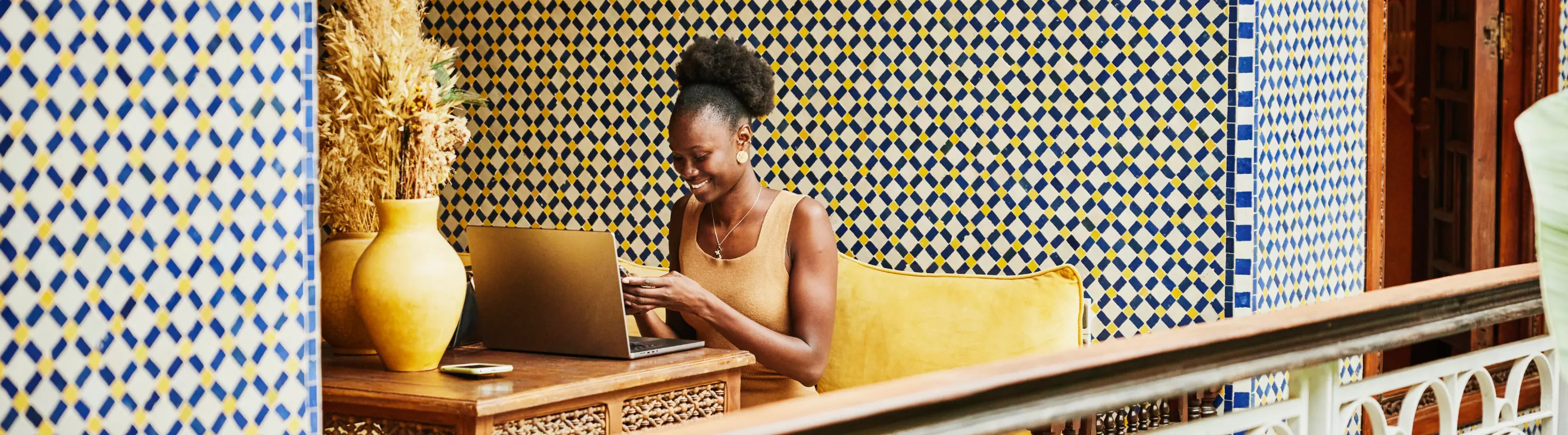 Young woman with travel insurance for digital nomads smiling at her phone while on a laptop on a balcony
