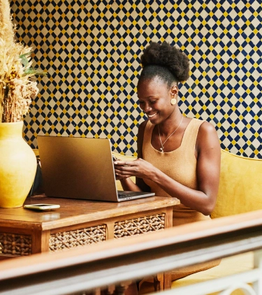 Young woman with travel insurance for digital nomads smiling at her phone while on a laptop on a balcony