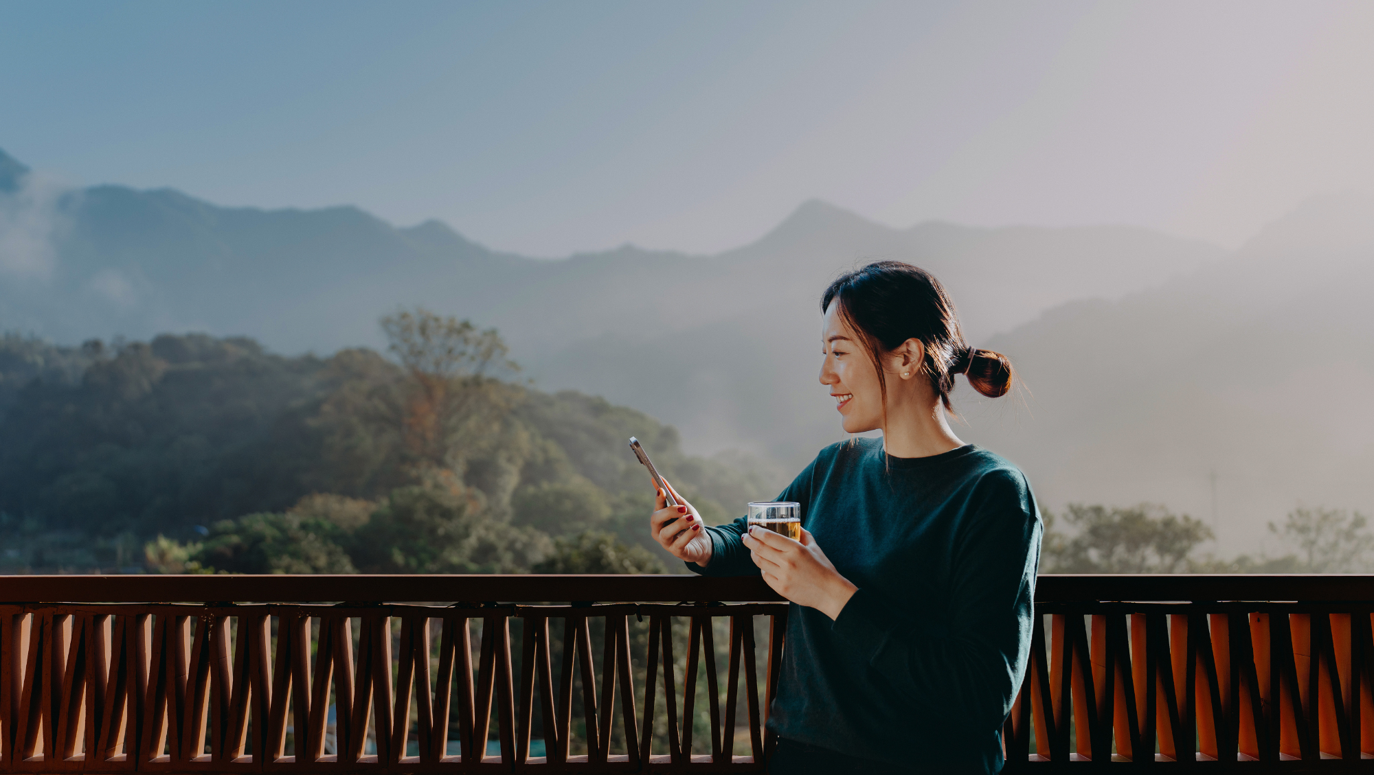 a woman looks at a travel app on her smartphone on a balcony with mountains in the background