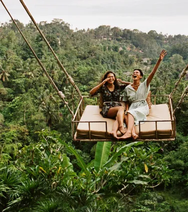 two women laugh on a giant jungle swing, insured with travel insurance for nature trips 