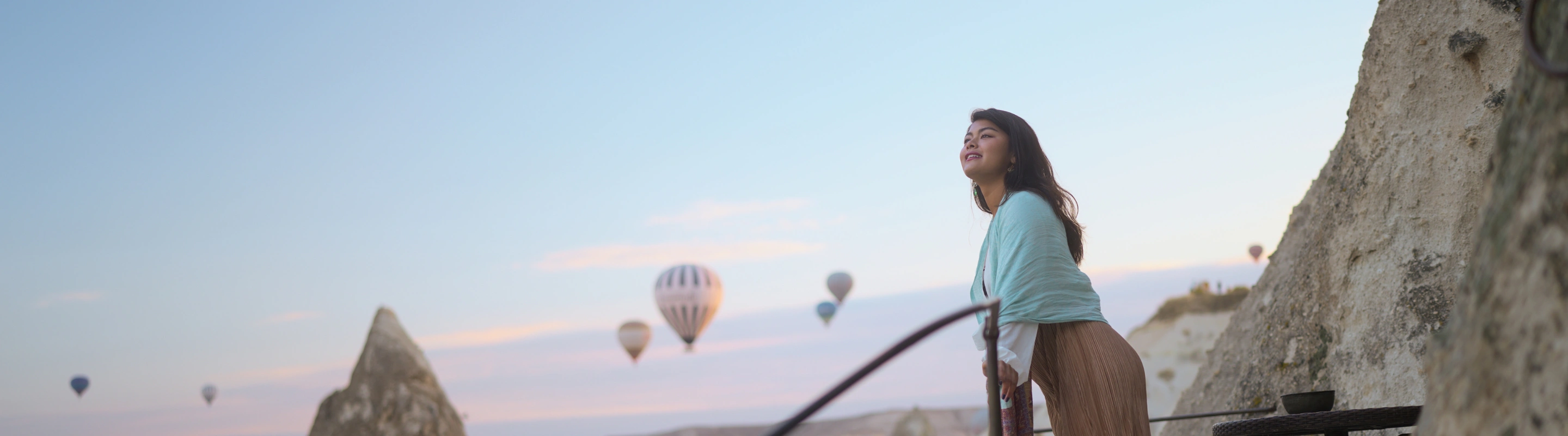 a young woman with international travel insurance leans on a rock ledge and takes in views of hot air balloons in Turkey