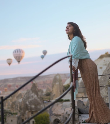 a young woman with international travel insurance leans on a rock ledge and takes in views of hot air balloons in Turkey