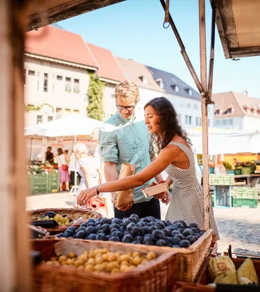 a young couple shops at a local market for fruit, insured with travel insurance for Germany