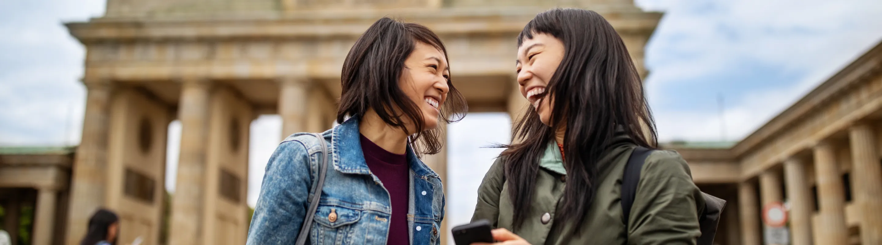 two female friends laugh in front of the Brandenburg Gate in Berlin, insured with travel insurance for Germany