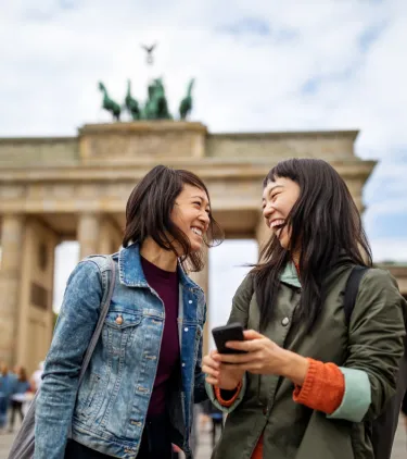 two female friends laugh in front of the Brandenburg Gate in Berlin, insured with travel insurance for Germany