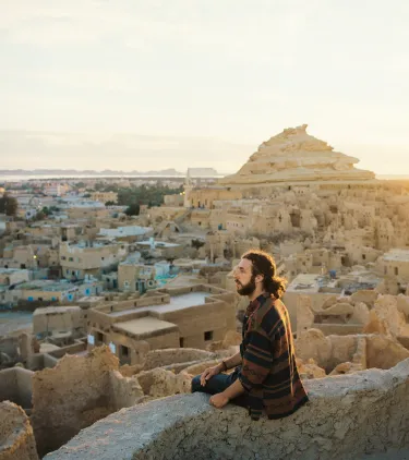 a young man sits on a ledge and overlooks the Siwa Oasis at sunset, insured with travel insurance for Egypt