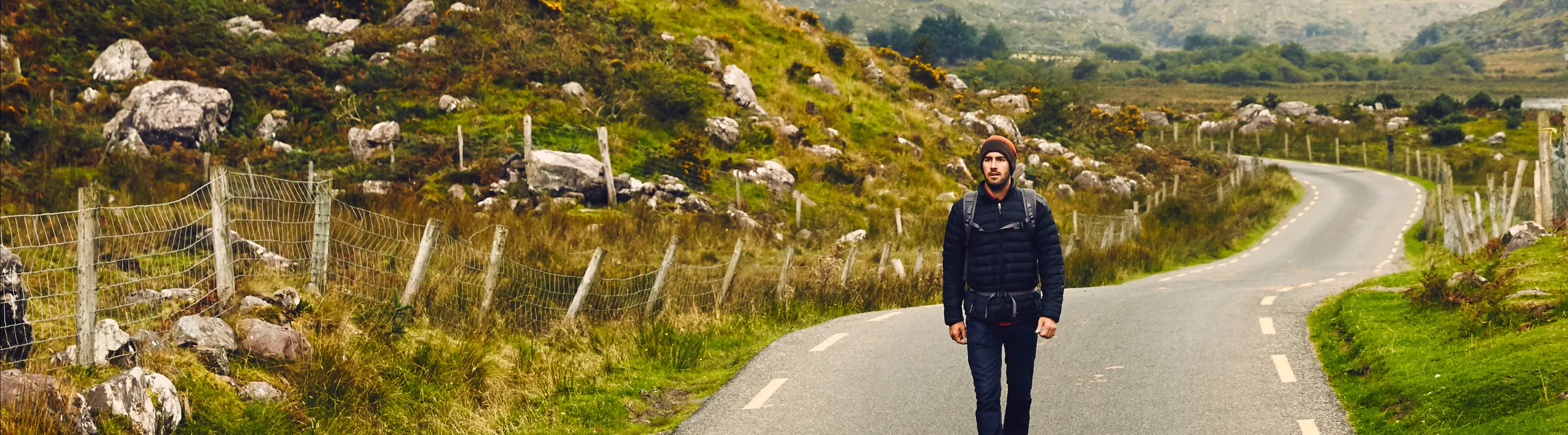 a young man with Ireland travel insurance walks down a scenic road near greenery and mountains 