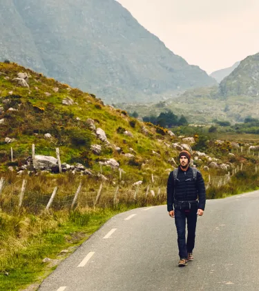 a young man with Ireland travel insurance walks down a scenic road near greenery and mountains 