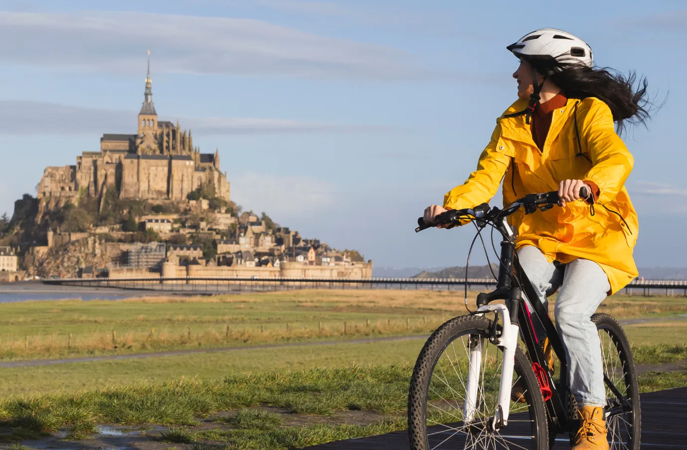 a woman in a yellow jacket rides her bike near Mont Saint Michel, insured with travel insurance for France