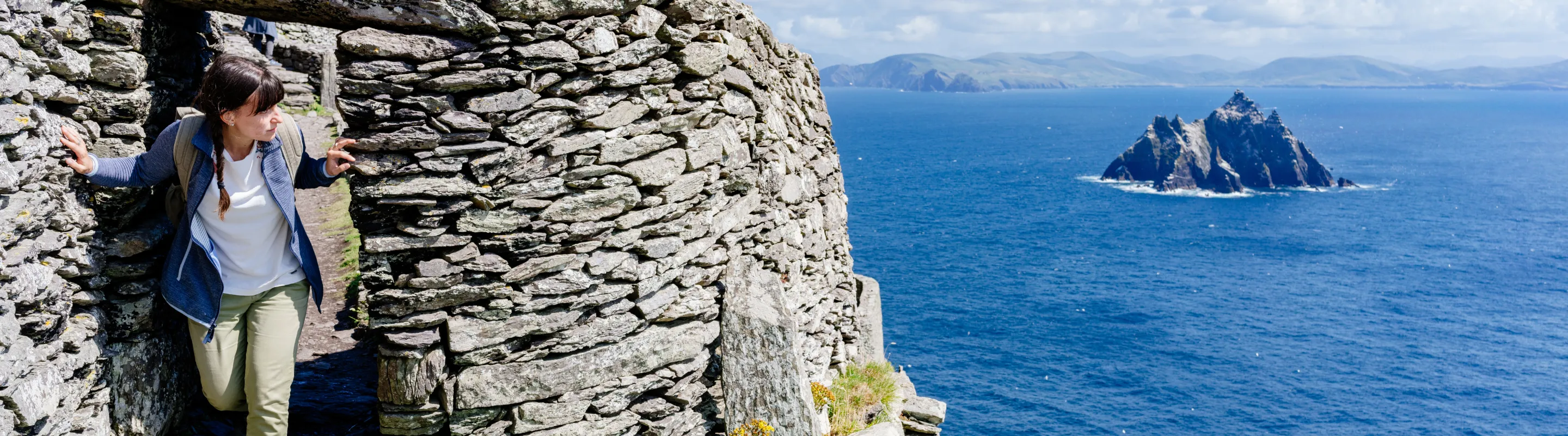 a young adult female hikes through the Skellig Michael ruins by the ocean, insured with travel insurance for Ireland