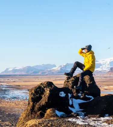 a man in a yellow jacket overlooks a black sand beach, insured with travel insurance for Iceland 
