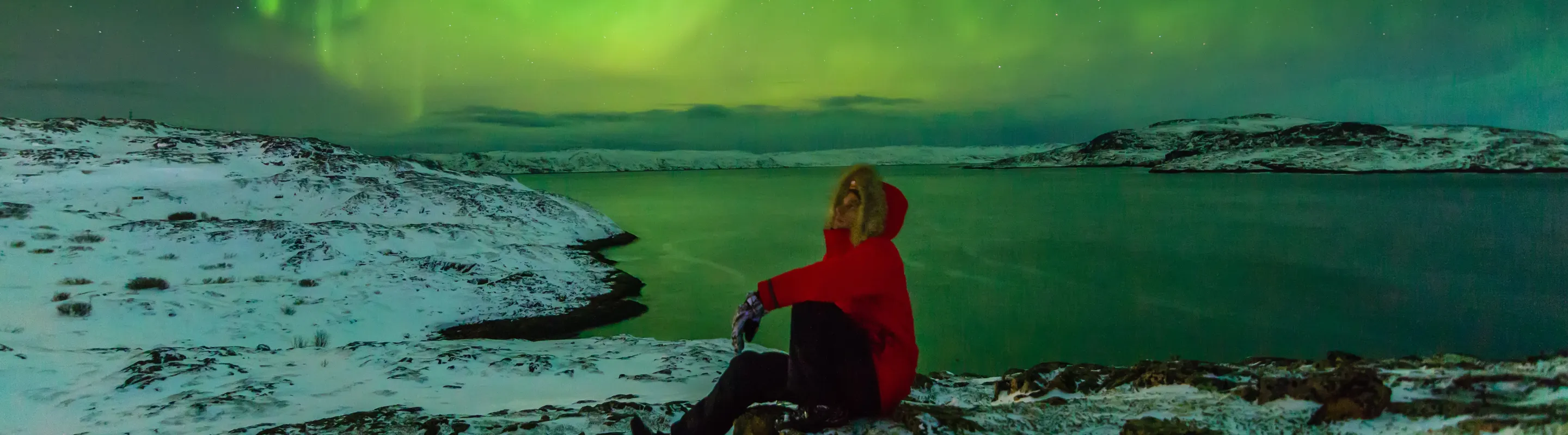 a traveler in a red jacket sits on the ground and watches the Northern Lights, insured with travel insurance for Iceland 