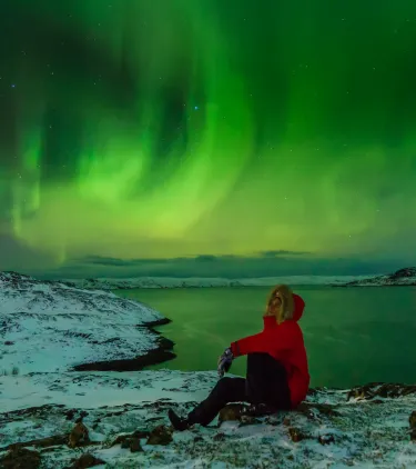 a traveler in a red jacket sits on the ground and watches the Northern Lights, insured with travel insurance for Iceland 