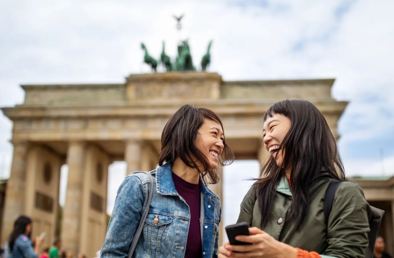 two female friends laugh in front of the Brandenburg Gate in Berlin, insured with travel insurance for Europe