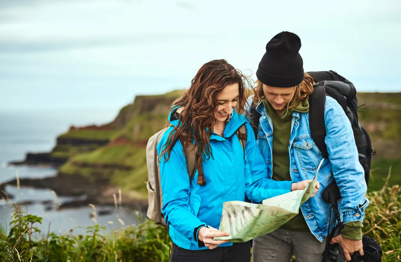 a young couple in blue jackets looks at a map while hiking along the coast, insured with travel insurance for Ireland 