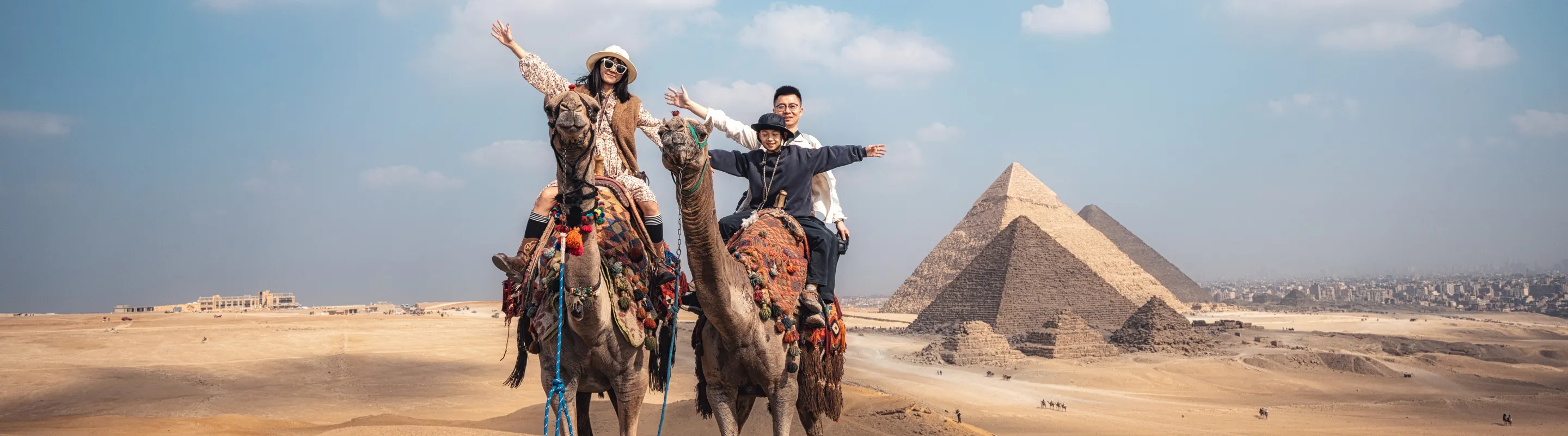 a family of three raises their arms as they ride camels at the Pyramids of Giza, insured with travel insurance for Egypt