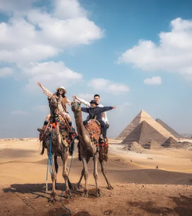 a family of three raises their arms as they ride camels at the Pyramids of Giza, insured with travel insurance for Egypt
